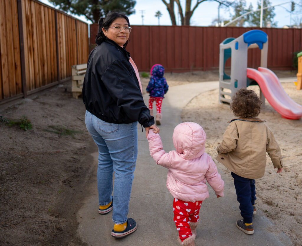 A woman leads two children around a walkway next to some playground equipment. The children's faces are not visible.