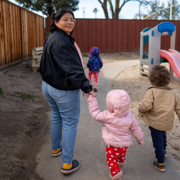 Una mujer guía a dos niños por un sendero junto a algunos juegos infantiles. Los rostros de los niños no son visibles.