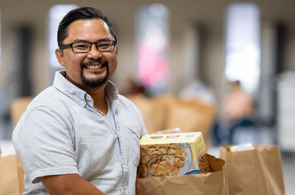 A Food Bank employee smiles at the camera next to a grocery bag with cereal and other food items.