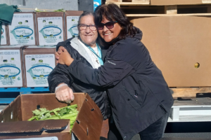 Two woman hug next to a bin of produce.
