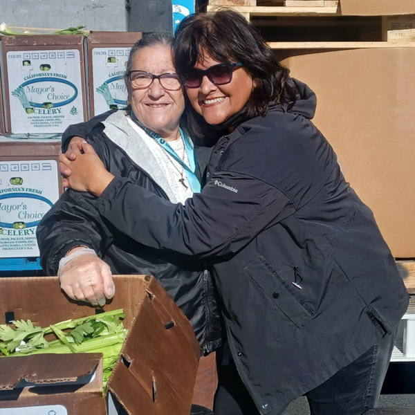 Two woman hug next to a bin of produce.