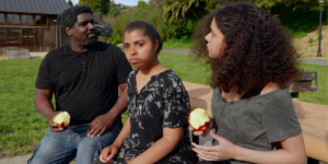 A father and his two teenage daughters eat apples in the park.